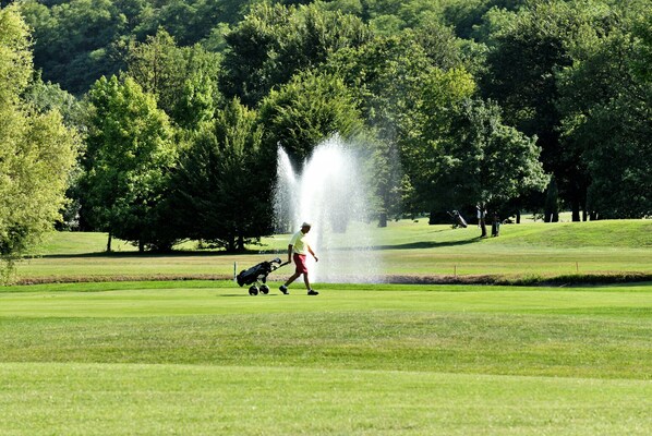 Campo de golf con sistema de riego, una persona empujando un carrito de golf y árboles al fondo.