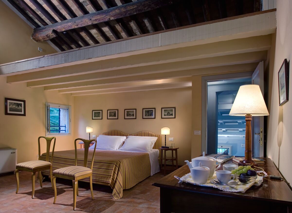 A hotel room with a bed, two chairs, a desk with a lamp, a window, and framed pictures on the wall.