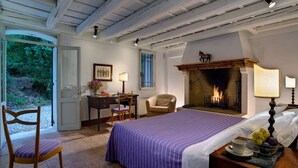 A bedroom with a bed, a desk, a chair, a fireplace, and a view of greenery.