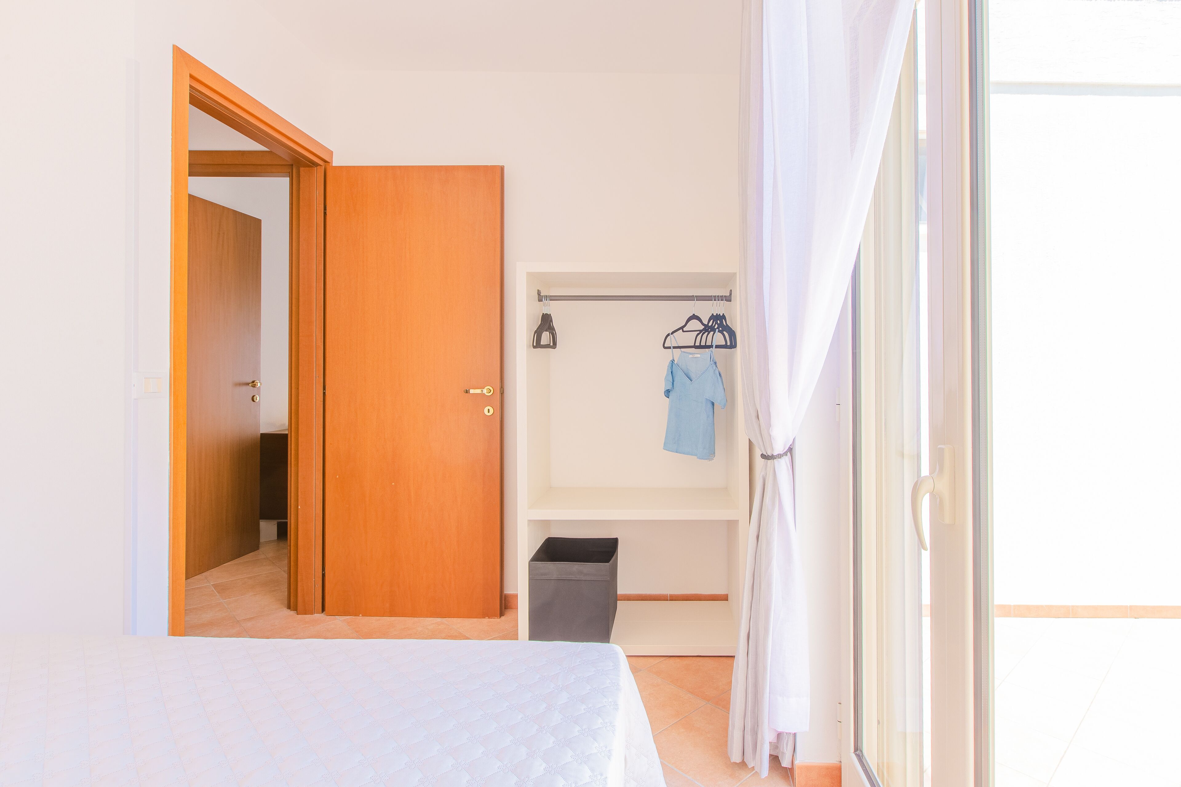 A bedroom with a bed, a wardrobe, a hanging rack, and a trash bin.
