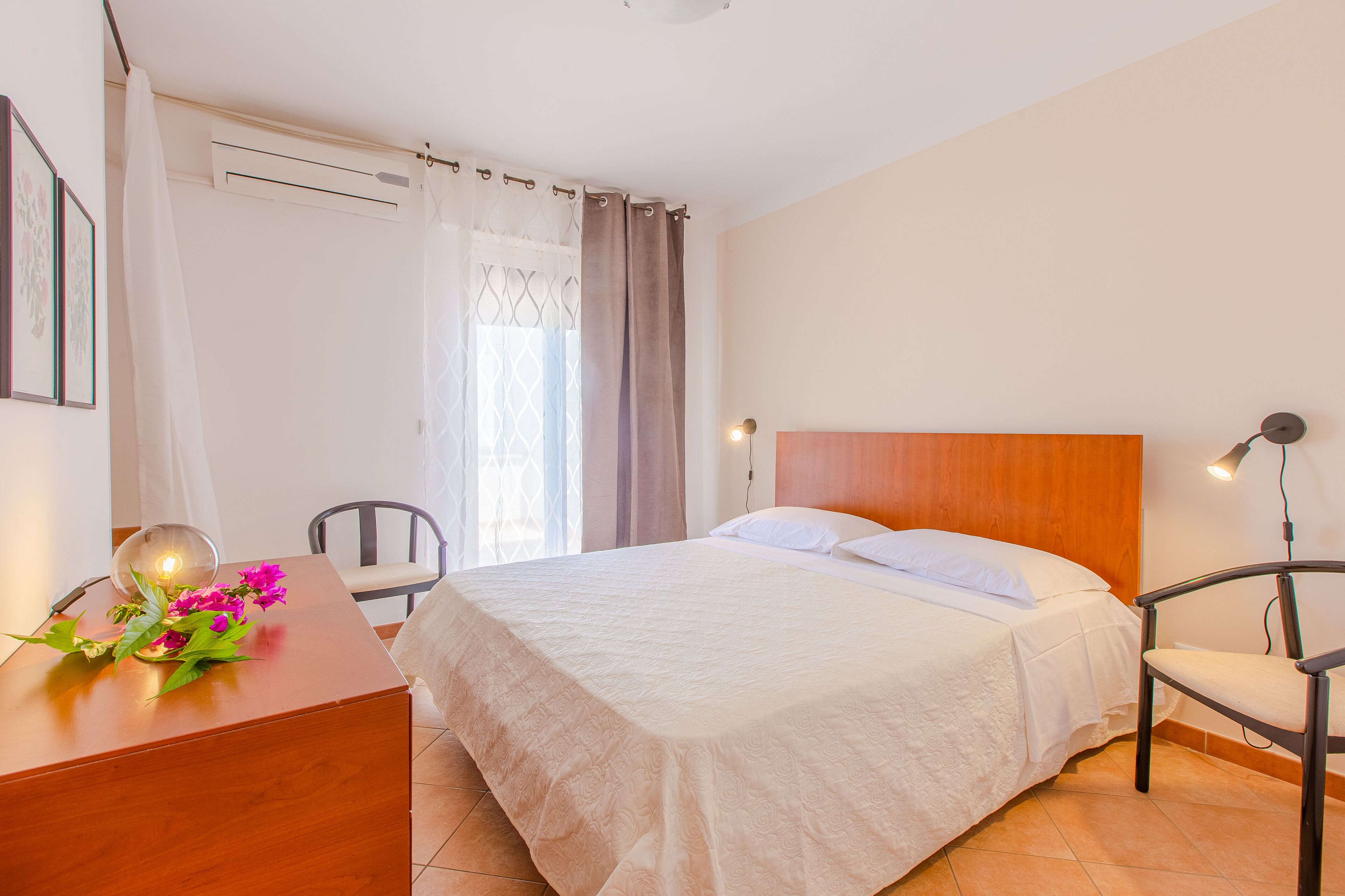 Una camera da letto ordinata con un letto, un comodino, una lampada, una sedia e una scrivania con un vaso di fiori.