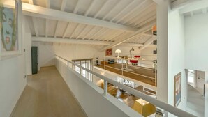A spacious loft-style room with a high ceiling and exposed beams, a balcony with a railing, and a view of the lower level.