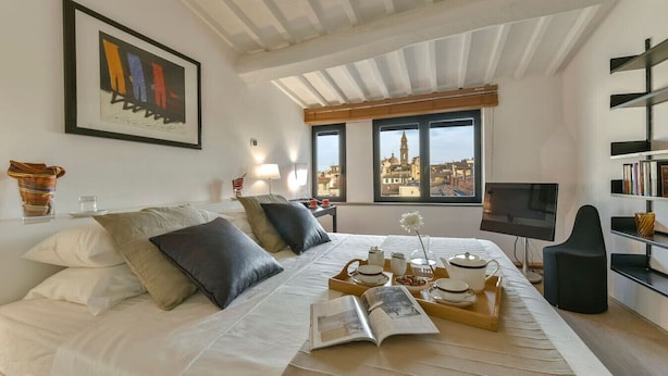 A modern hotel room with a large bed, a TV, a bookshelf, and a view of a cityscape through the windows.