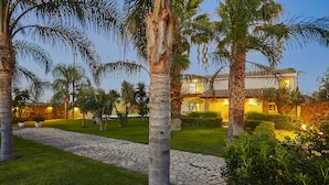 A well-maintained garden with palm trees, a stone pathway, and a building with warm lighting. - Villa with splendid pool near the beaches, 4 bedrooms (Marinella di Selinunte)