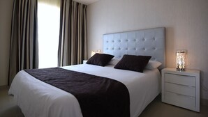 A hotel room with a large bed, white bedding, a tufted headboard, a bedside table with a lamp, and curtains on the window. - Apartment  WITH SEA VIEWS and excelent location - IDEAL FOR FAMILIES (Costa Calma)