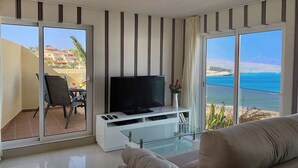 A modern living room with a flat-screen TV, a glass coffee table, and a sofa. The room has large sliding glass doors leading to a balcony with a view of the ocean and a mountain. - Apartment  WITH SEA VIEWS and excelent location - IDEAL FOR FAMILIES (Costa Calma)