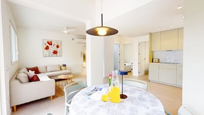 Un salon moderne comprenant un canapé, une table à manger recouverte d’une nappe blanche, un luminaire suspendu et un coin cuisine avec des placards et un plan de travail.