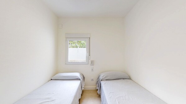 A small, narrow room with two single beds, a window with a view of greenery, and a white door.