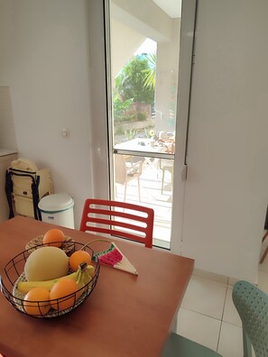 Un coin repas avec une table dressée pour deux personnes, un panier de fruits et une vue sur la terrasse extérieure.