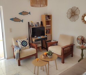 A living room with a sofa, armchair, coffee table, TV, bookshelf, decorative fish wall art, and a fan.
