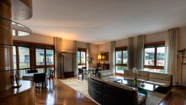 A spacious living room with a curved black sofa, a glass coffee table, and a dining area with a table and chairs. Large windows provide natural light and views of the outside.