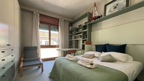 A hotel room with a bed, a chair, a small table, a bookshelf, and a view of the outside balcony.
