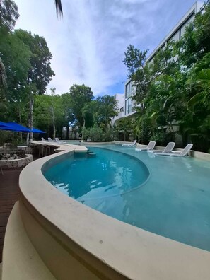 A curved swimming pool with lounge chairs, surrounded by trees and a building with large windows.