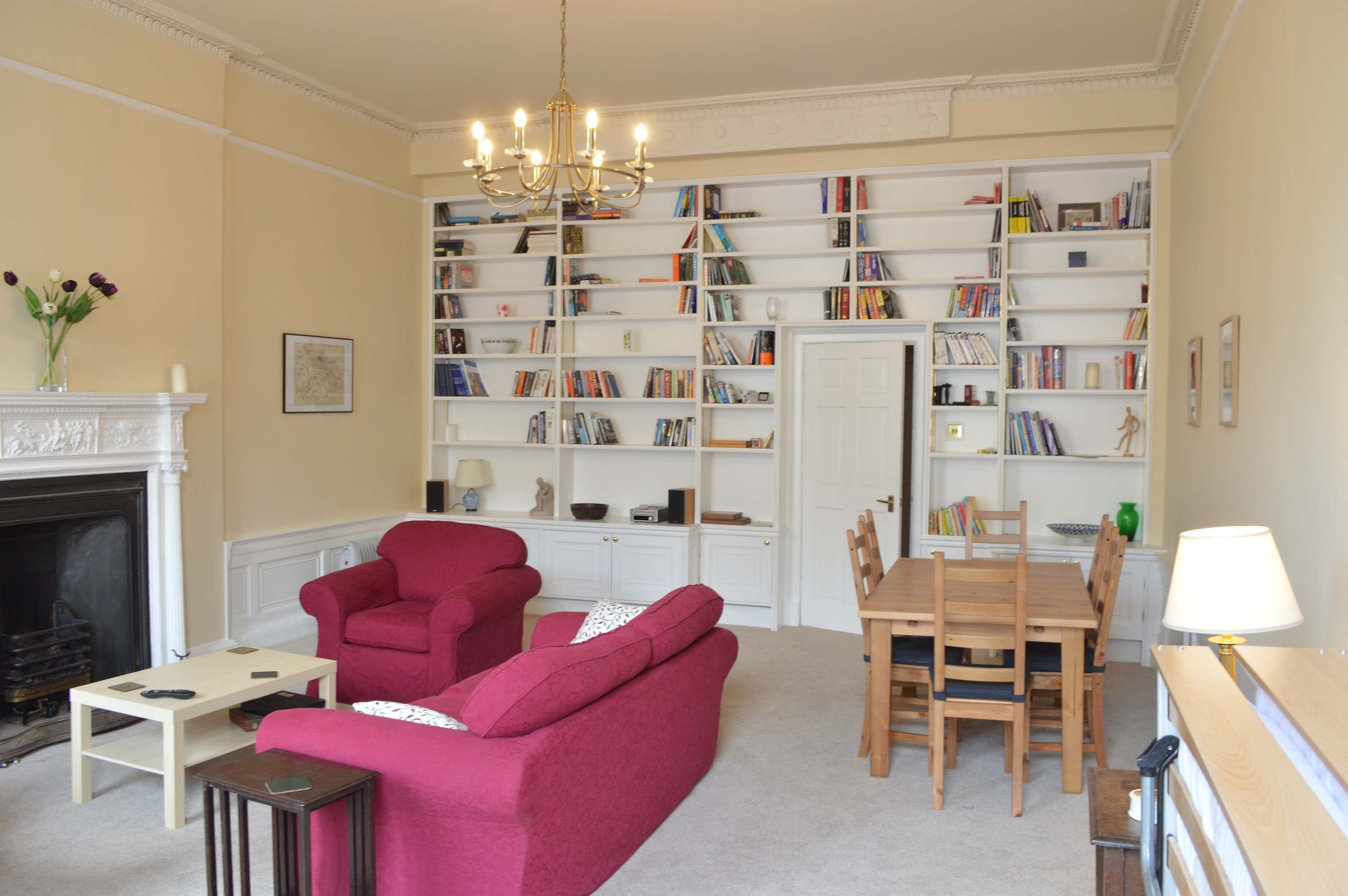 A living room with a fireplace, a red sofa, a wooden dining table with chairs, and a bookshelf filled with books.