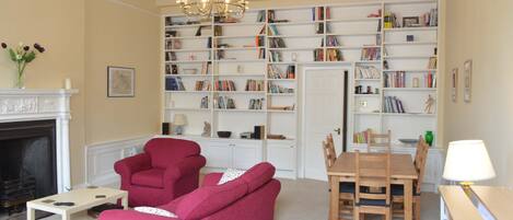A living room with a fireplace, a red sofa, a wooden dining table with chairs, and a bookshelf filled with books.