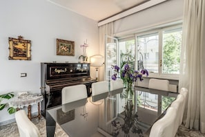 A dining room with a glass table, white chairs, a piano, and framed artwork on the wall.