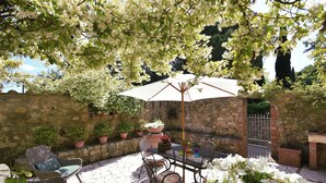 An outdoor patio with wicker furniture, a table, and a large umbrella. There are potted plants and a stone wall in the background.