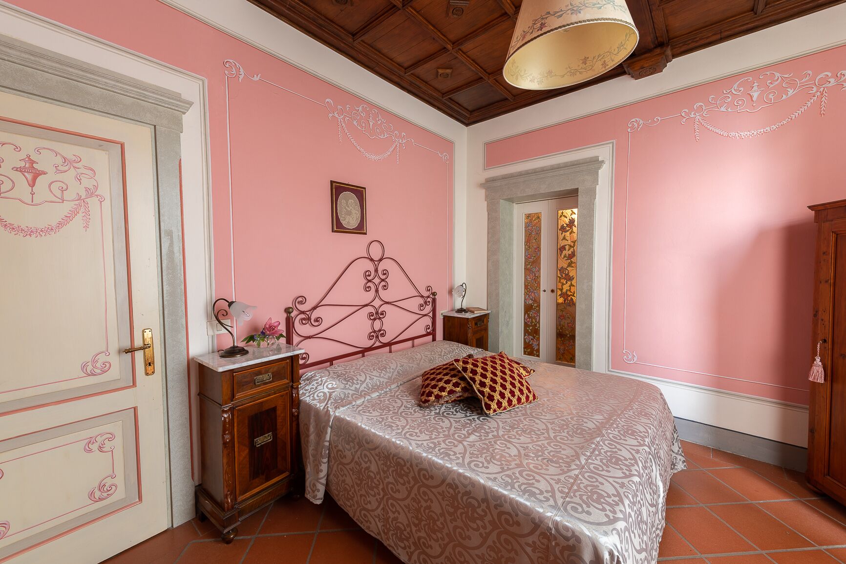 A bedroom with pink walls, a bed with patterned bedding, a nightstand, and a wooden wardrobe.