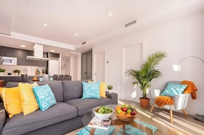 A modern living room with a grey sofa, a dining area, a kitchen, and a potted plant.