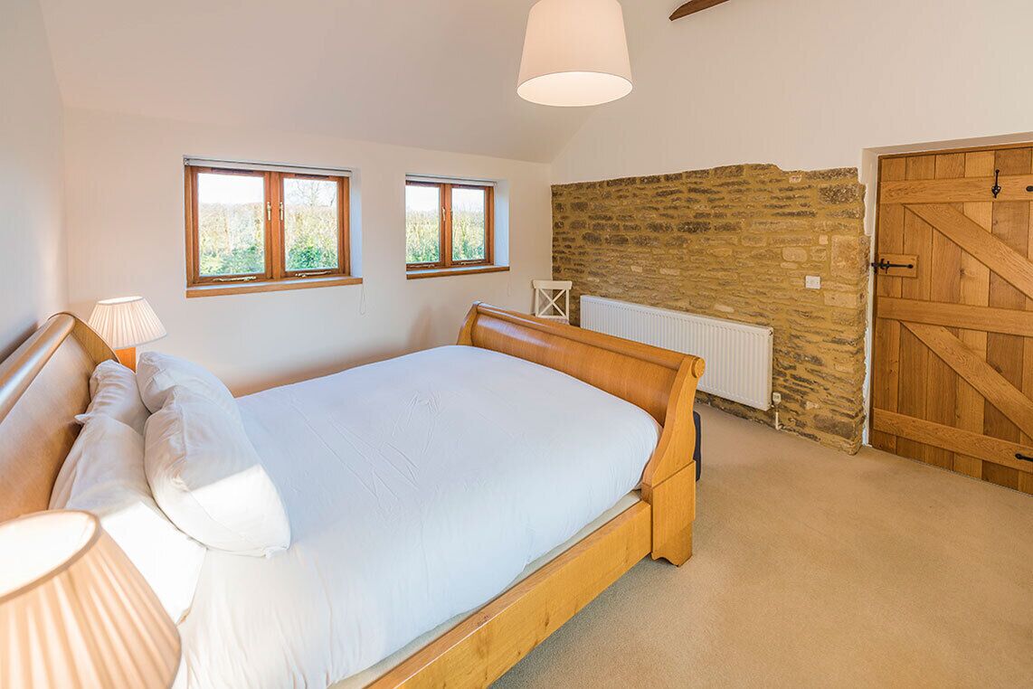 A bedroom with a wooden bed, two windows, a stone wall, a radiator, and a wooden door.