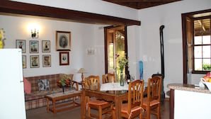 Comedor con mesa de madera, sillas y un centro de mesa floral. Se observa un sofá con funda estampada de cuadros al fondo.