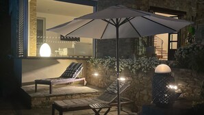 Une terrasse en pierre avec des chaises longues et un parasol, un mur en pierre et un intérieur bien éclairé, visible à travers une grande fenêtre.