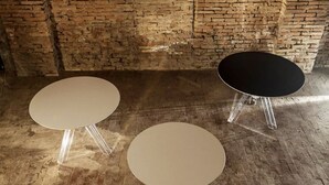 Trois tables rondes aux pieds transparents, dans une pièce dont un mur est en briques.