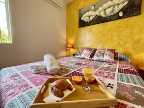 Un lit bien fait, recouvert d’une courtepointe, une tablette avec des croissants et des tranches d’orange, un verre de jus d’orange et un tableau accroché au mur.