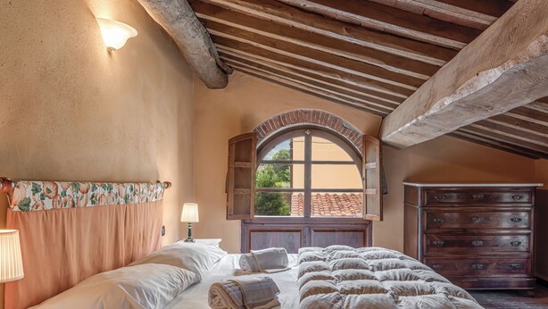 A bedroom with a large bed, a wooden dresser, a window with an arched top, and a lamp on the nightstand.