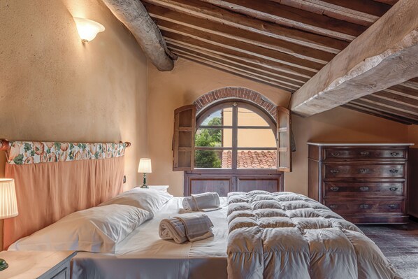 A bedroom with a large bed, a wooden dresser, a window with an arched top, and a lamp on the nightstand.