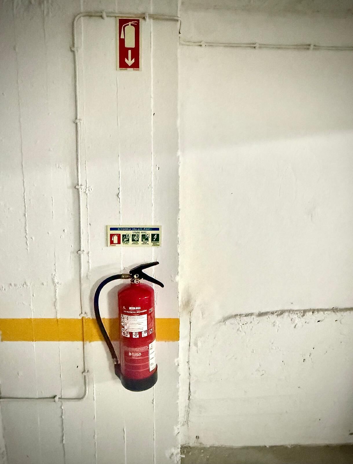 Estintore fissato a una parete bianca con una striscia gialla, un segnale di uscita di emergenza rosso e un cartello di sicurezza verde.