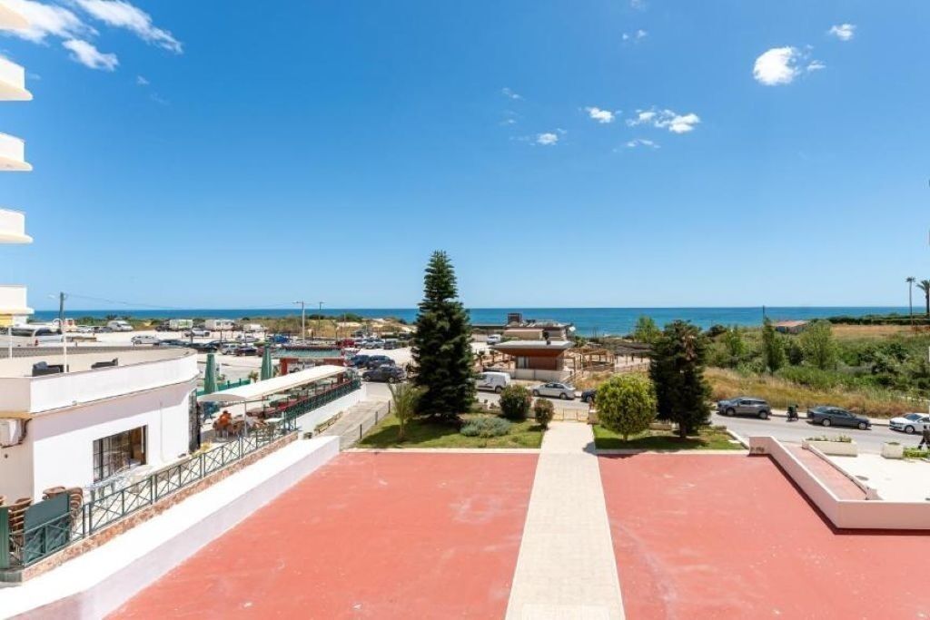 Eine Dachterrasse mit rotem Boden, einem zentralen Weg und Blick auf das Meer sowie Gebäude im Hintergrund.