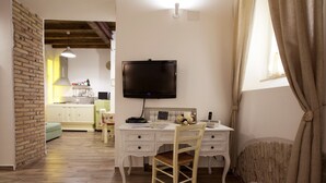 A room with a wooden floor, a white desk with a chair, a television mounted on the wall, and a window with curtains.
