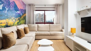 Ein modernes Wohnzimmer mit einem beigen Sofa, zwei weißen Beistelltischen, einem Flachbildfernseher und einem großen Fenster mit Vorhängen.