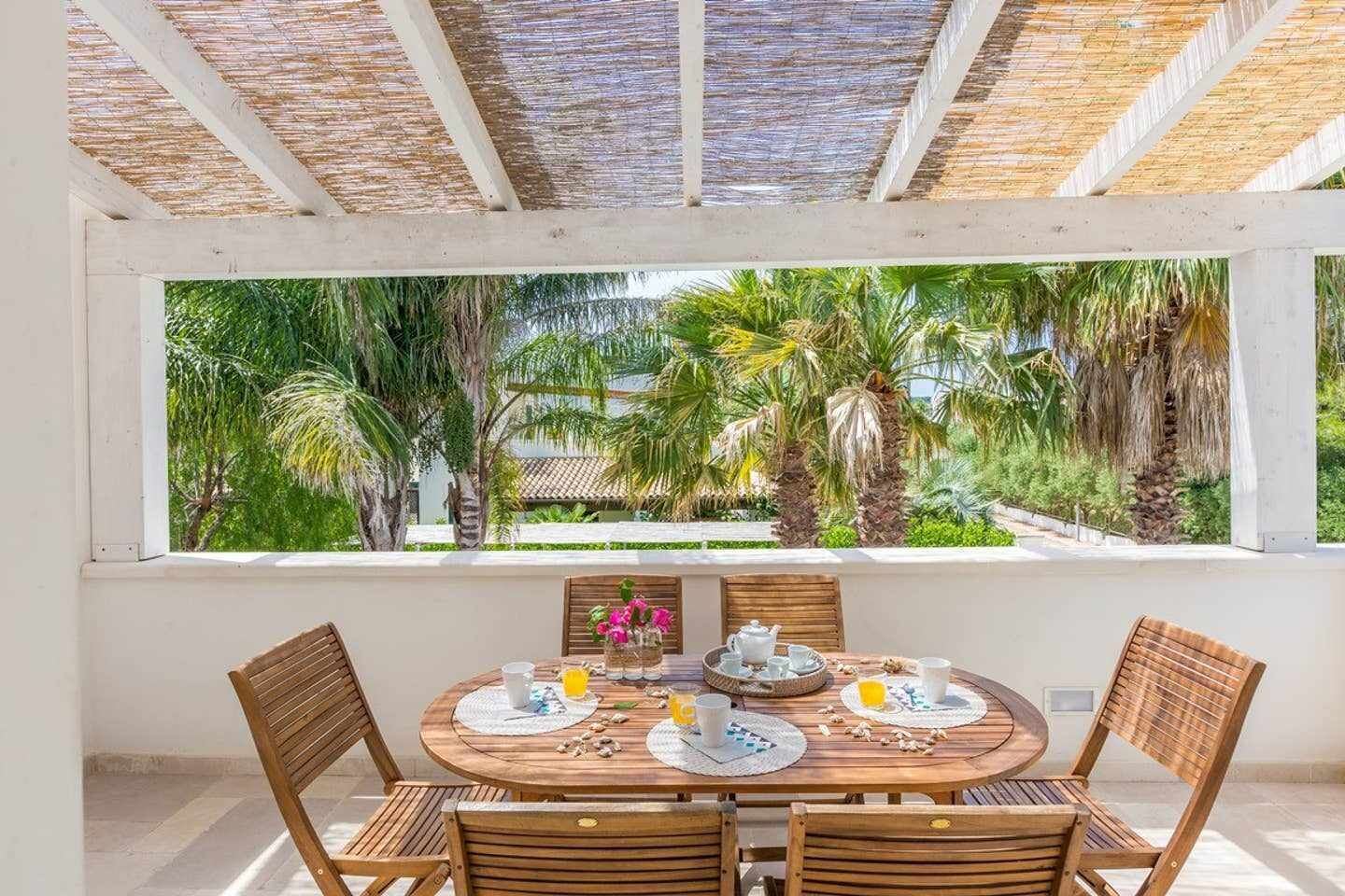 A round wooden table with four chairs, a window with a view of palm trees, and a thatched roof.