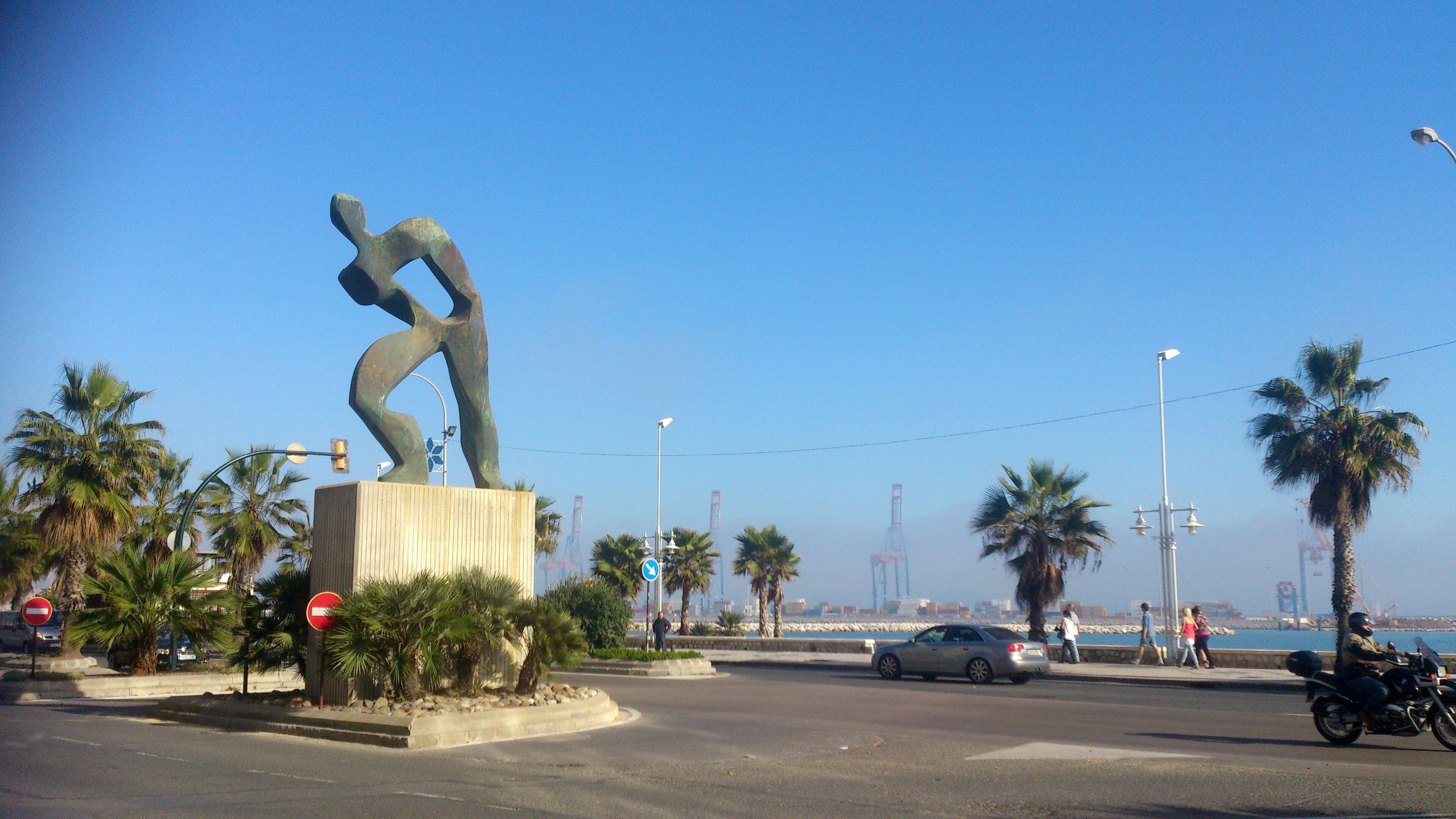 A sculpture in a public square with palm trees and a clear sky.