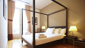 A four-poster bed with white linens, two bedside tables with lamps, a chair, and a view through a glass door.