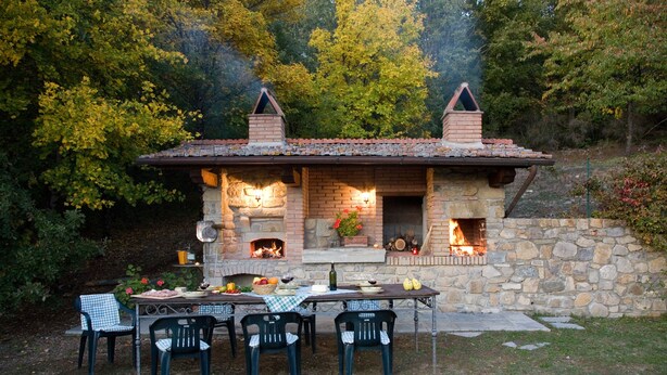 A stone building with a tiled roof, a fireplace, and outdoor seating with chairs and a table. - Ancient country villa with pool in the hills of Tuscany, surrounded by woods  (Poppi)