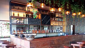 Zona de bar con mostrador y taburetes de madera, estanterías con botellas, luces colgantes y pared decorada.