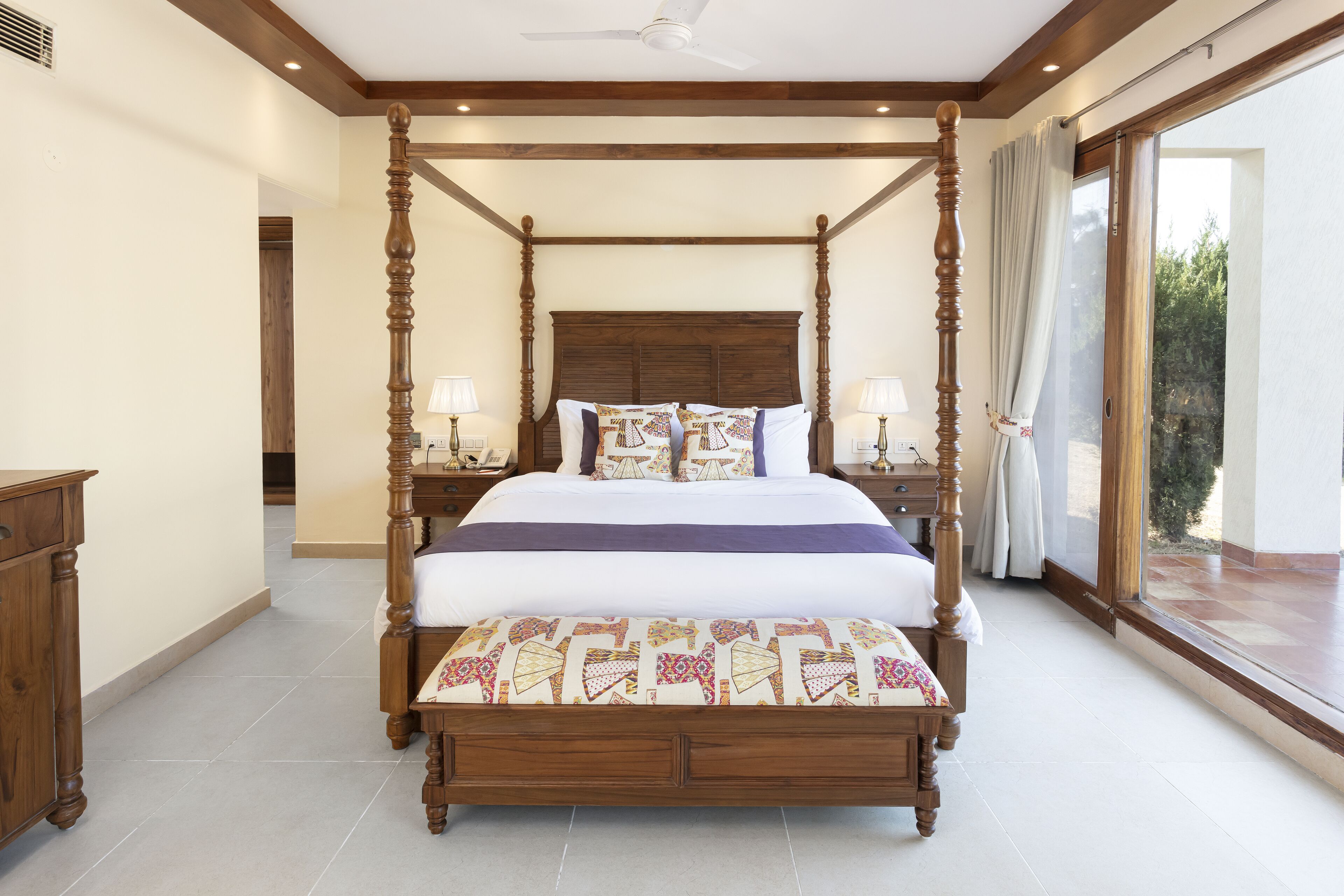 A four-poster bed with a wooden headboard, two bedside tables with lamps, and a view of the outdoors through a large window.