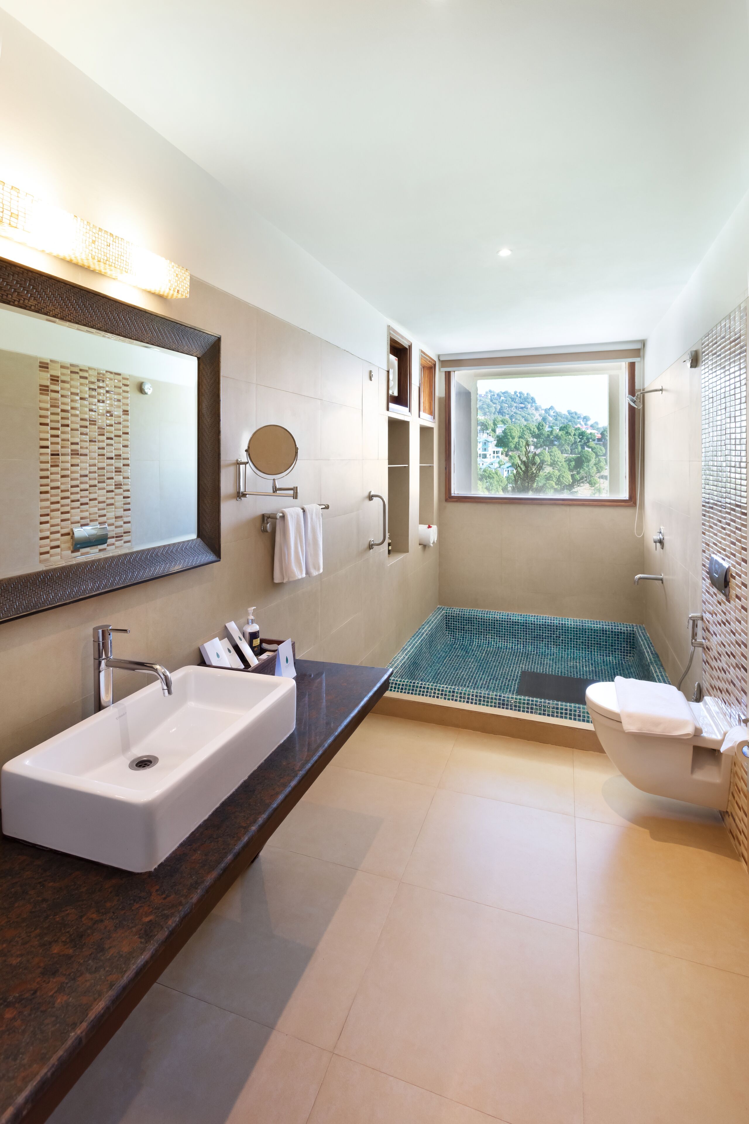 A modern bathroom with a large mirror, a bathtub with a green mosaic floor, a toilet, and a vanity with a sink.