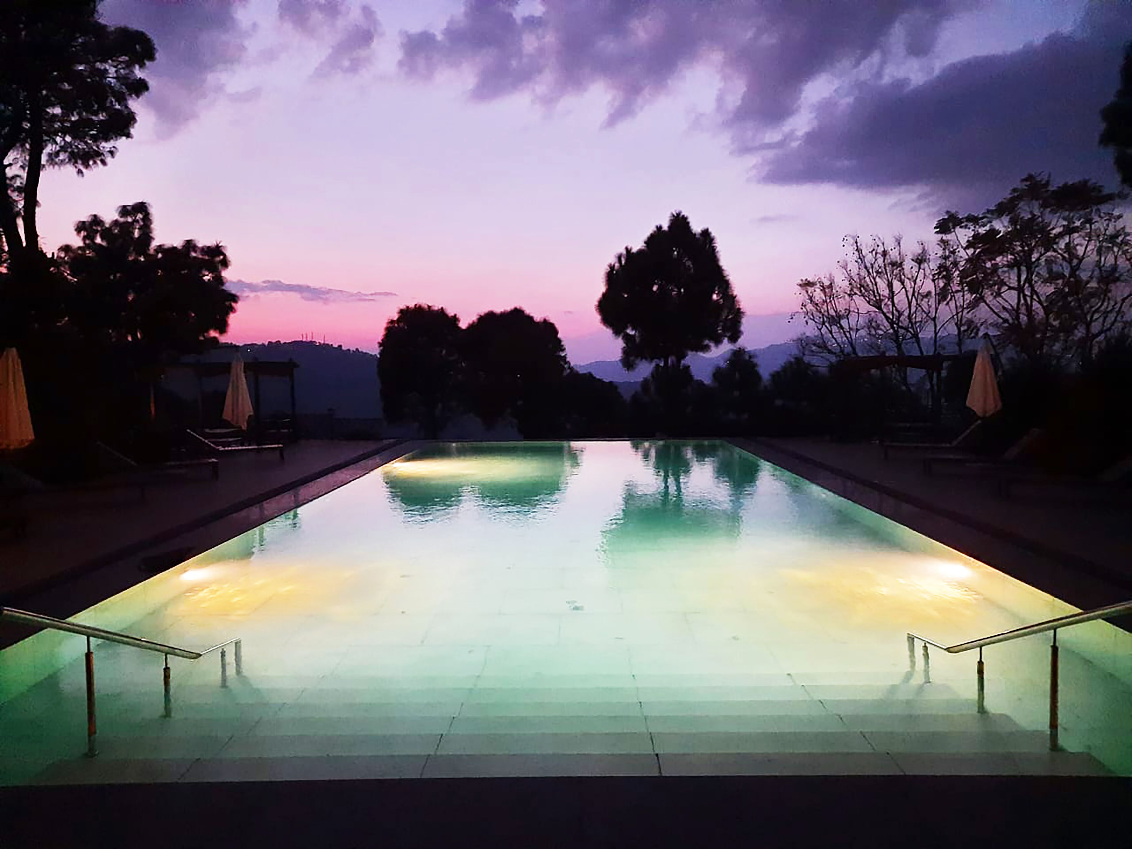 A rectangular swimming pool with a clear water surface, surrounded by lounge chairs and umbrellas, set against a twilight sky.