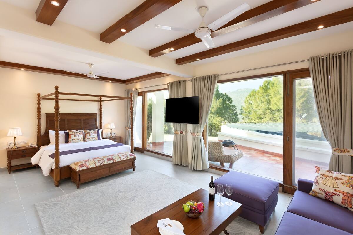 A spacious bedroom with a four-poster bed, a flat-screen TV, and a balcony with a view of the outdoors.