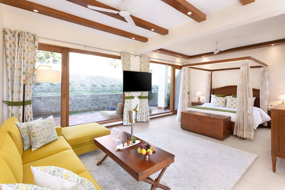 A spacious bedroom with a four-poster bed, a yellow sofa, a flat-screen TV, and a balcony with curtains.