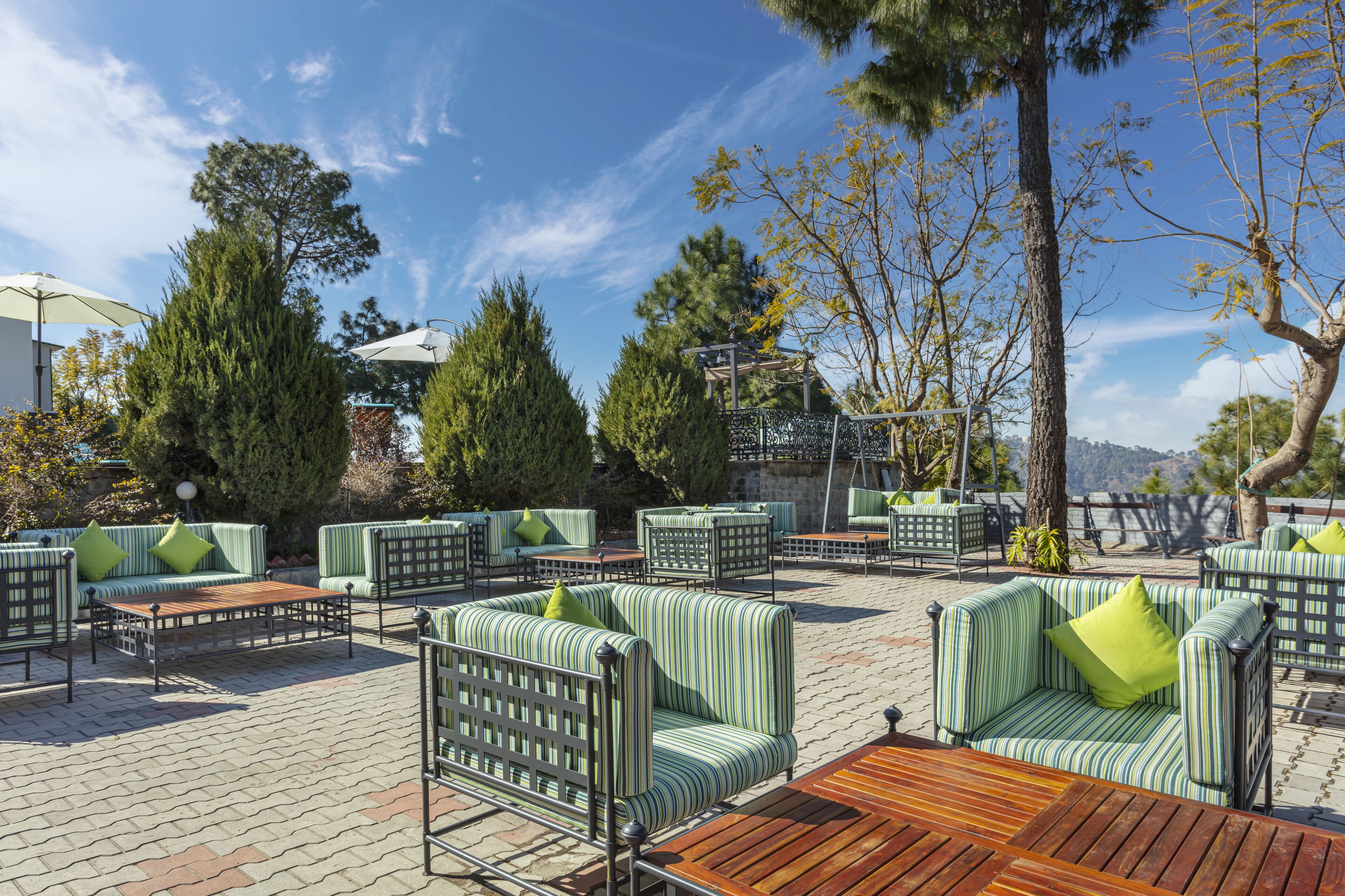 An outdoor patio area with striped green sofas and tables, surrounded by trees and a clear sky.