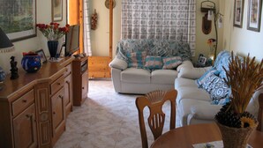 A cozy living room with a sofa, a dining table, and a TV. There is a wooden cabinet and a vase with flowers on the table. - Apartment in private urbanization with pool and garden area in Santa Pola (Santa Pola)