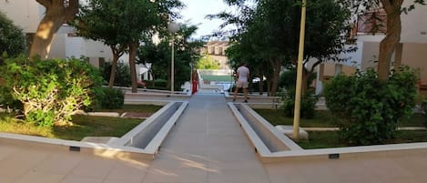 A pathway flanked by trees and bushes, leading to a building with a white facade.