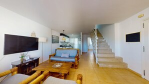 A spacious living area with a sofa, wooden coffee table, and a TV mounted on the wall. A staircase leads to an upper floor. - Maravilloso Ático a tan solo 150 m de la playa. Wifi. A/A. 5 pers. (Dénia)
