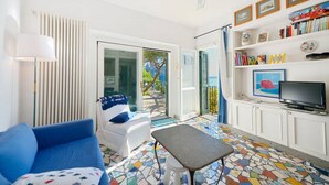 A living room with a blue sofa, a small table, a TV, and a bookshelf with books and decorative items. - Cottage by the Sea in Cartaromana Bay, with a View of Aragonses Castle  (Ischia)