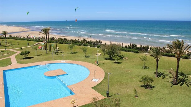 A pool with a view of the beach and ocean, surrounded by palm trees and a clear sky.